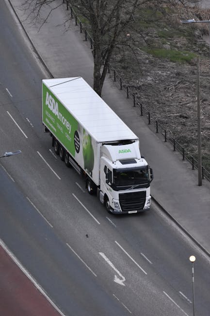 An aerial view of a white moving truck with the ASDA Money logo parked on the side of a city street, adjacent to a pavement lined with a metal fence and sparse trees. The truck is positioned on a mult