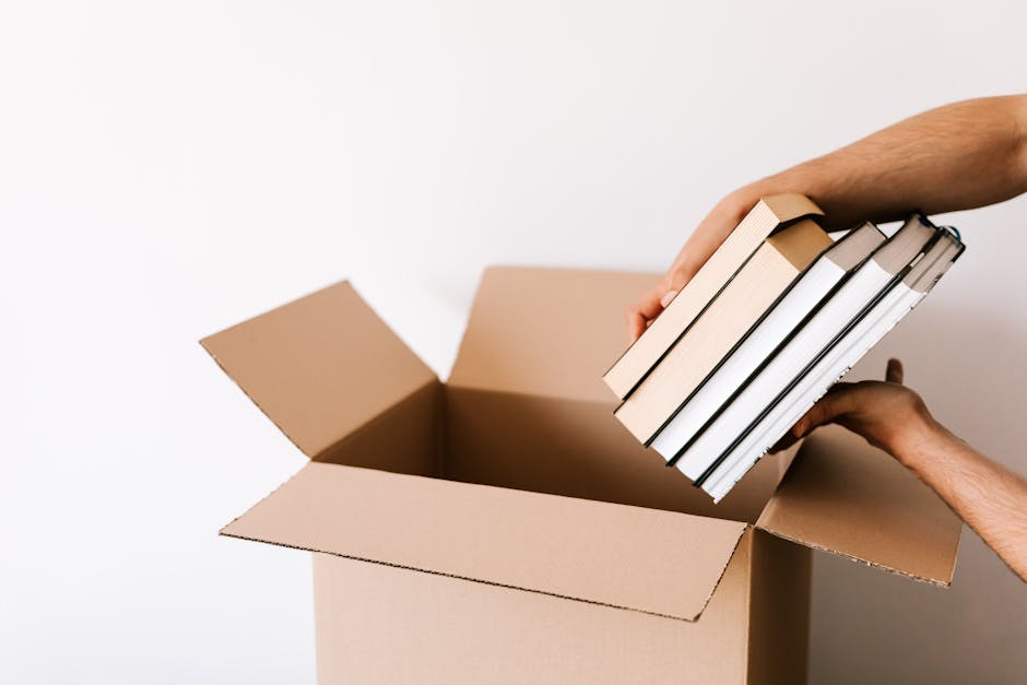 A person with dark hair and a beard, wearing a brown t-shirt, is seen placing a closed, orange hardcover book into a cardboard box during a home relocation process. The box is partially filled with va