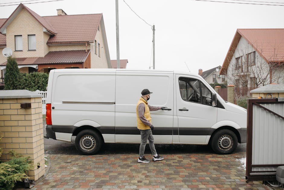 A man wearing a black cap, grey trousers, a yellow and grey jacket, and a face mask is opening the sliding door of a white commercial van during a home relocation process. The van is parked on a brick