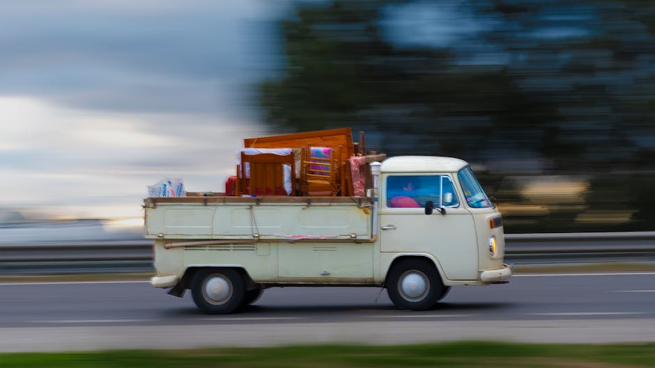 A man wearing a black cap, grey trousers, a yellow and grey jacket, and a face mask is opening the sliding door of a white commercial van during a home relocation process. The van is parked on a brick