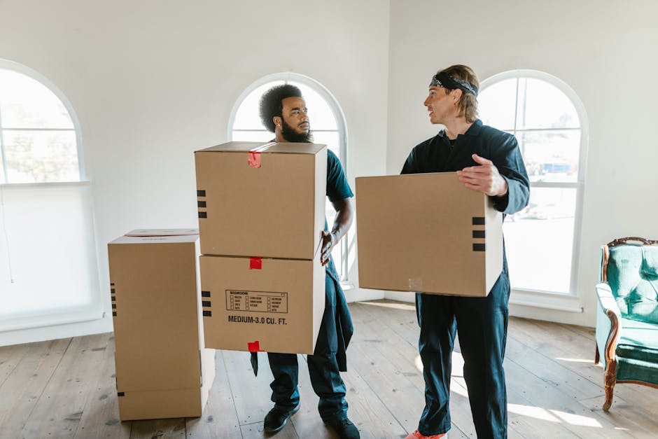 Two professional movers from Man and a Van Merton are inside a well-lit room with white walls and large arched windows, engaged in a home relocation process. The mover on the left, with curly hair and facial hair, is holding two medium-sized cardboard moving boxes stacked vertically, with red and black tape securing them. The mover on the right, wearing a headband and dressed in dark casual clothing, is carrying a single cardboard box while gesturing as if giving instructions or coordinating the loading process. In the background, there are additional packed boxes placed on the wooden floor near the corner, and a green upholstered armchair with wooden accents is positioned against the wall. Natural light enters through the windows, illuminating the scene, which depicts the packing and furniture transport phase of domestic removals, emphasizing the logistics involved in a house move managed by [COMPANY_NAME], focused on efficient packing and moving services.