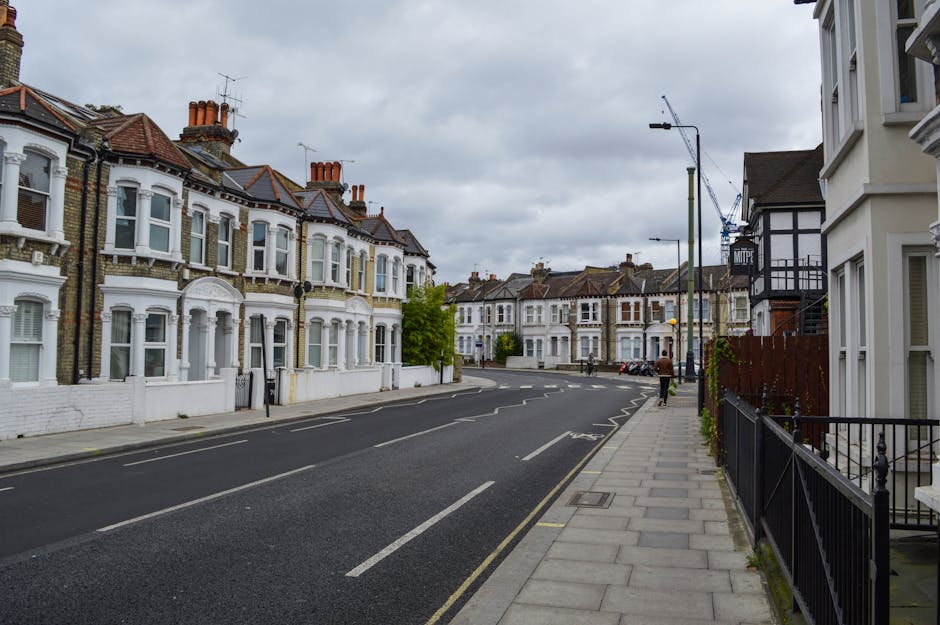 An empty residential street on Wimbledon Park Road lined with Victorian-style terraced houses featuring bay windows, brick and white-painted facades, and chimneys under a cloudy sky. The pavement on the right side has a black metal fence alongside residential steps, while on the left side, the road has marked parking bays and a series of parked cars. The street is illuminated by a tall streetlight and is currently quiet with no moving vehicles or pedestrians. The image depicts an urban environment suitable for home relocation or furniture transport, with no visible moving equipment or boxes in this scene but naturally aligning with the context of house removals services provided by Man and a Van Merton.