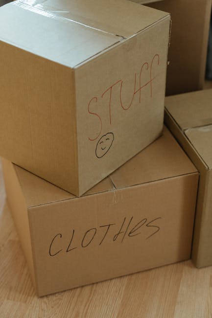 A man with dark, curly hair and a beard is indoors, holding a medium-sized cardboard box labeled 'OFFICE' in black text, with additional labels indicating its contents and packaging specifications. Th